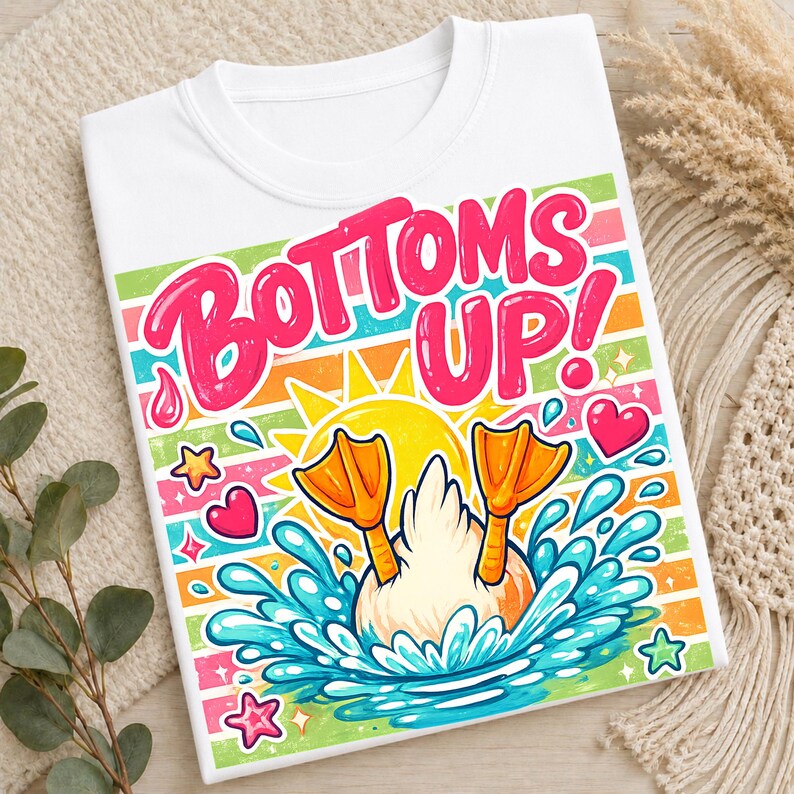 May include: White t-shirt with a colorful graphic featuring a duck diving into water, with the words "Bottoms Up!" in pink. The design includes a sun, hearts, and stars.
