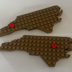 May include: Two brown, textured, North Carolina state-shaped objects with red heart accents. The objects are covered in small, circular studs. One heart is near the top, and the other is near the bottom of the state outline.