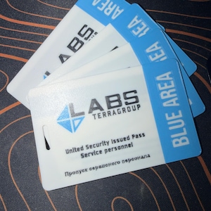 Op de afbeelding: Een stapel rechthoekige witte plastic identificatiebadges met een blauwe verticale streep aan de rechterkant met de tekst BLUE AREA. De badges tonen het logo LABS TERRAGROUP, de tekst United Security Issued Pass, Service personnel.