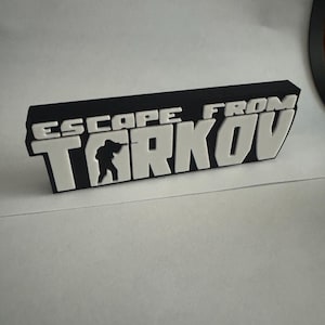 May include: A black and white sign with the text "ESCAPE FROM TARKOV". The sign has a 3D effect, with the letters and silhouette of a person in white against a black background. The sign is rectangular and appears to be made of a solid material.