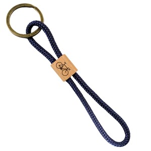 May include: A navy blue keychain with a brass ring and a wooden accent. The wooden accent features a bicycle graphic. The keychain is made of a woven rope material.