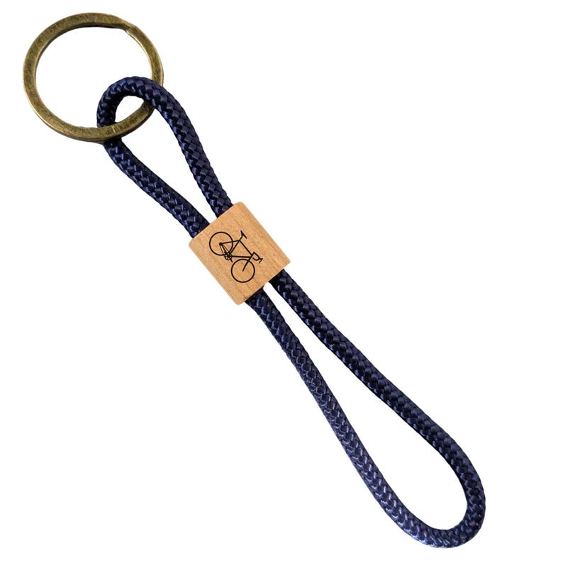 May include: A navy blue keychain with a brass ring and a wooden accent. The wooden accent features a bicycle graphic. The keychain is made of a woven rope material.
