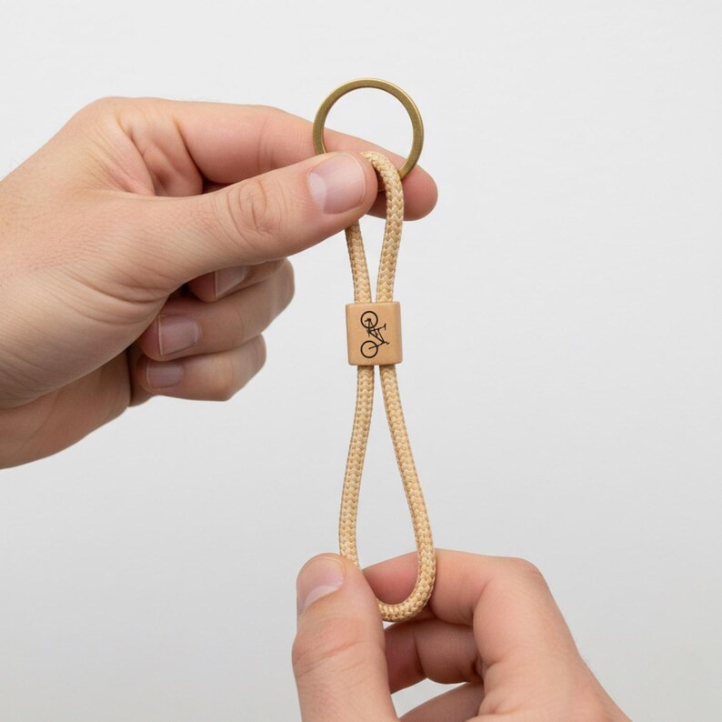 May include: A beige rope keychain with a brass ring and a wooden bead featuring a bicycle graphic. The keychain is held between two hands against a white background. The rope is looped.