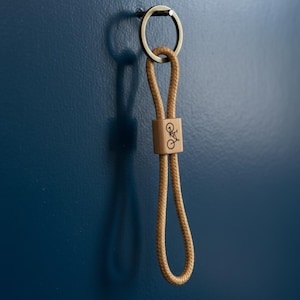 May include: A tan rope keychain with a wooden accent featuring a bicycle graphic. The keychain has a metal ring and is hanging against a dark blue wall. The wooden accent is rectangular.