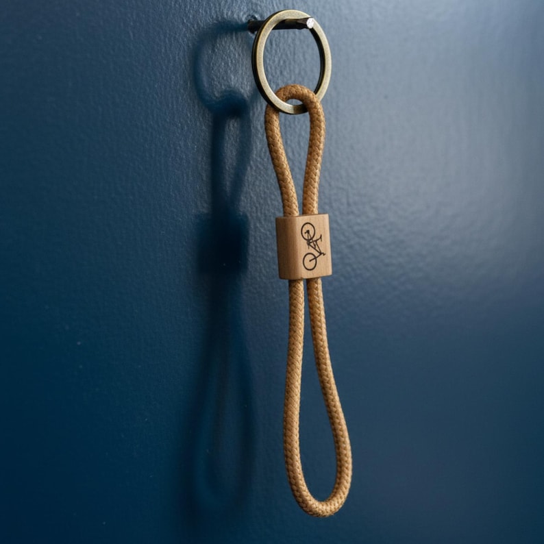 May include: A tan rope keychain with a wooden accent featuring a bicycle graphic. The keychain has a metal ring and is hanging against a dark blue wall. The wooden accent is rectangular.