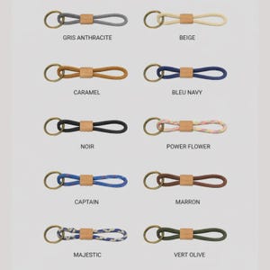 May include: A collection of keychains in various colors, each featuring a loop of rope connected to a wooden accent and a brass-colored ring. Colors include gray, beige, caramel, navy, black, and floral patterns. Text labels identify each color.