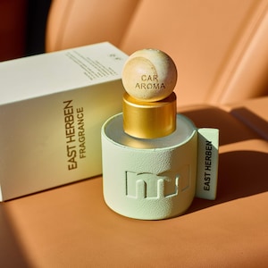 May include: A car air freshener with a light green cylindrical base and a wooden sphere top, labeled "CAR AROMA." The product is next to a white box with the text "EAST HERBEN FRAGRANCE." The items are on a tan surface.