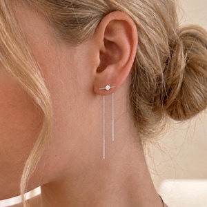 May include: A silver threader earring featuring a small circular stud connected to two delicate chains of varying lengths that hang down from the earlobe. The earring is crafted from a polished silver-toned metal with a minimalist design.