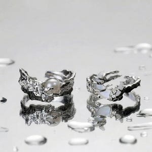 May include: Two silver-toned rings with an organic, textured design. One ring features a pearl, the other has a white, iridescent stone. The rings are set against a reflective surface with water droplets.
