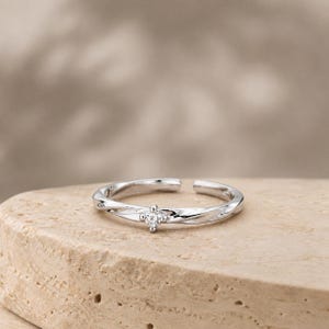 May include: A silver-toned open ring featuring a polished, twisted band design. A single, small, clear, round-cut gemstone is set in a four-prong setting at the center of the band, which remains open at the back to allow for adjustable sizing.