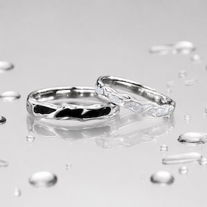 以下が含まれることがあります： 不規則で有機的なバンドデザインのシルバー調の指輪2点。一方の指輪はテクスチャー加工された金属の中に滑らかで光沢のある黒いインレイが施され、もう一方の指輪はバンドに沿ってきらめく白いグリッターのようなインレイが施されています。