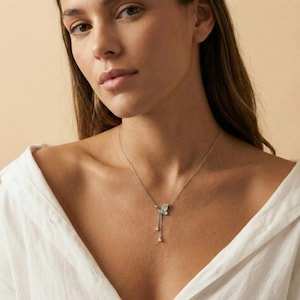 May include: A silver necklace featuring a butterfly pendant with two dangling teardrop charms. The butterfly has light blue accents. The necklace is worn on a person wearing a white shirt.