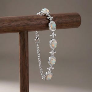 Peut inclure: Un bracelet en argent orné de pierres d'opale ovales, chacune entourée de petits gemmes transparents. Le bracelet est doté d'une fine chaîne et d'un fermoir. Les opales présentent un aspect blanc laiteux et irisé. Le bracelet est présenté sur un support en bois.