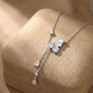 May include: A silver necklace featuring a butterfly pendant with a pearlescent center and sparkling accents. The necklace has a delicate chain with dangling teardrop and round-cut gems, creating an elegant and feminine design.