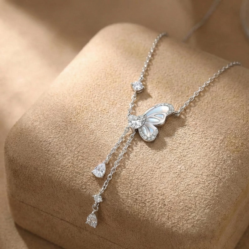 May include: A silver necklace featuring a butterfly pendant with a pearlescent center and sparkling accents. The necklace has a delicate chain with dangling teardrop and round-cut gems, creating an elegant and feminine design.