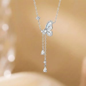May include: A silver necklace featuring a butterfly pendant with white enamel and sparkling accents. The butterfly has a dangling chain with three teardrop-shaped gems. The necklace is displayed against a blurred, warm-toned background.