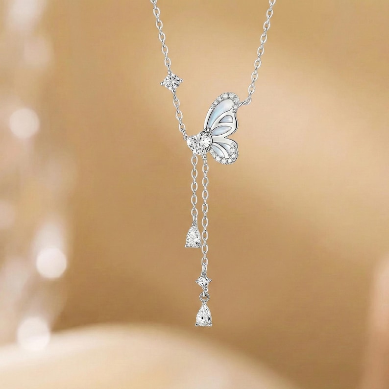May include: A silver necklace featuring a butterfly pendant with white enamel and sparkling accents. The butterfly has a dangling chain with three teardrop-shaped gems. The necklace is displayed against a blurred, warm-toned background.