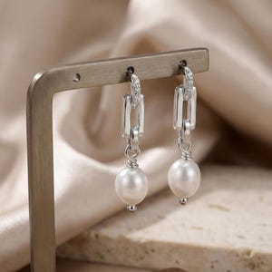 May include: A pair of silver earrings with a chain link design and a dangling white pearl. The top of the earring has a small row of clear stones. The earrings are displayed on a metal stand.