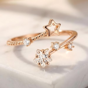 May include: Rose gold ring with star and floral detailing. The open design features a star-shaped cutout and is embellished with small, sparkling gemstones. The ring's band has a dotted texture, enhancing its delicate appearance.
