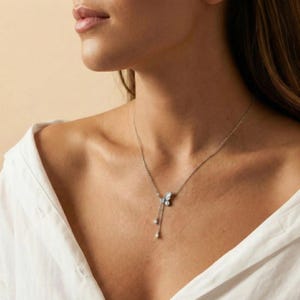 May include: A silver butterfly pendant necklace with dangling teardrop-shaped charms. The butterfly has intricate detailing and is adorned with small, sparkling accents. The necklace is worn against a white shirt.
