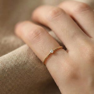 May include: A thin gold ring featuring a single small round clear gemstone set in the centre. The band has a delicate beaded texture along its surface, creating a refined appearance. The metal has a warm yellow tone and a polished finish.