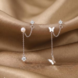 May include: A pair of silver earrings. One earring has a pearl and a dangling clear gemstone, connected by a chain. The other earring features a butterfly charm and a dangling butterfly, also connected by a chain. The earrings are set against a gold background.