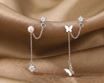 Op de afbeelding: Een paar zilveren oorbellen. Eén oorbel heeft een parel en een bungelende heldere edelsteen, verbonden door een ketting. De andere oorbel heeft een vlinderbedel en een bungelende vlinder, ook verbonden door een ketting. De oorbellen staan tegen een gouden achtergrond.