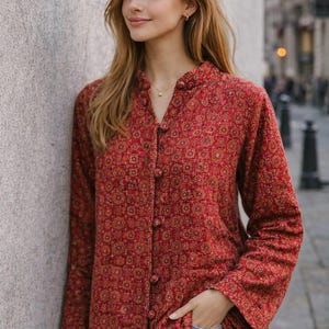 Puede incluir: Una chaqueta acolchada roja de manga larga con un patrón geométrico repetitivo en tonos naranja, amarillo y rojo oscuro. La chaqueta tiene un cuello mao, cierre frontal con botones cubiertos de tela y un dobladillo recto.