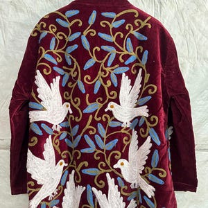 May include: A long-sleeved burgundy velvet coat featuring intricate embroidery of white doves and swirling gold vines with light blue leaves. The design covers the back of the garment, showcasing a symmetrical pattern of birds in flight amidst foliage.