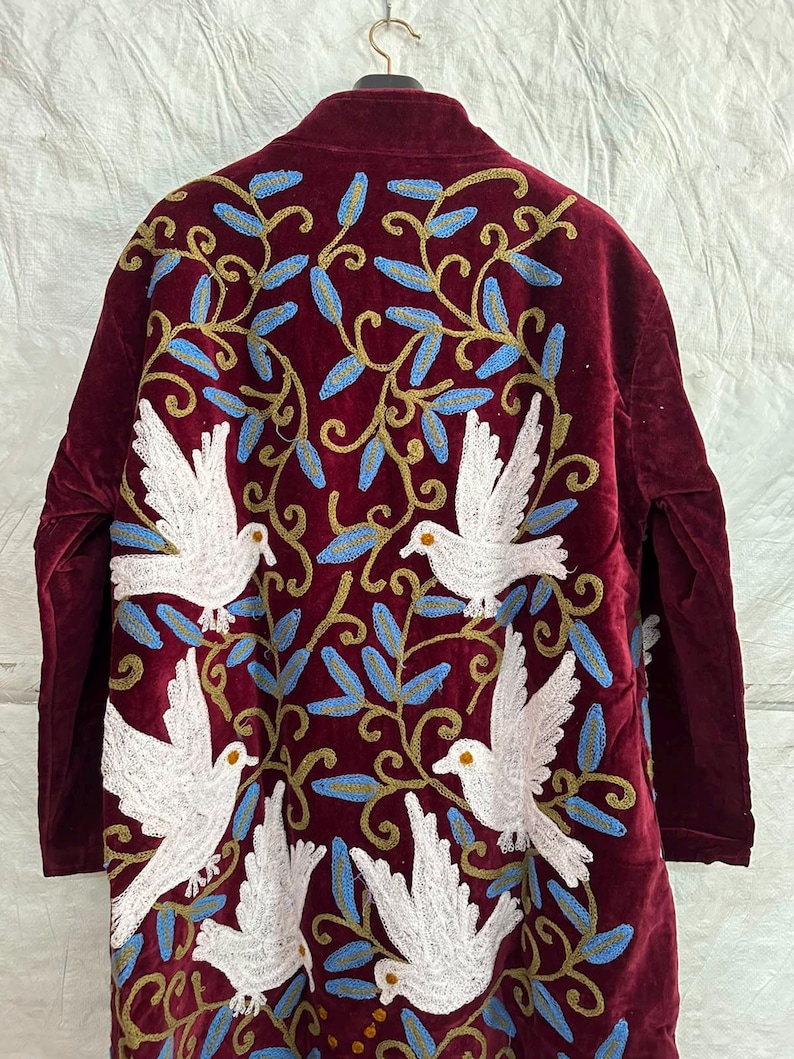 May include: A long-sleeved burgundy velvet coat featuring intricate embroidery of white doves and swirling gold vines with light blue leaves. The design covers the back of the garment, showcasing a symmetrical pattern of birds in flight amidst foliage.