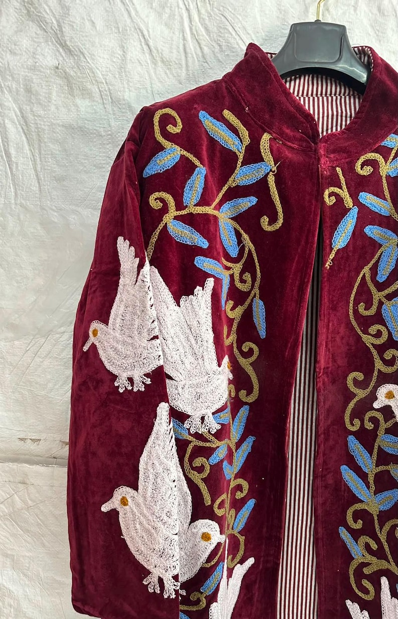 May include: A deep burgundy velvet jacket featuring intricate embroidery of white birds and gold vines with blue leaves. The garment has a high collar and a vertical red and white striped lining visible along the front opening, hanging on a black hanger.