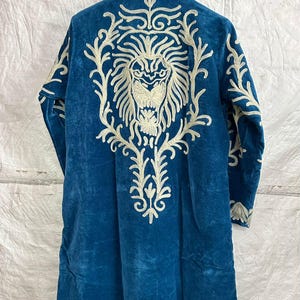 May include: A long-sleeved, deep blue velvet coat featuring intricate, cream-colored embroidery on the back. The design showcases a large, detailed lion head surrounded by ornate, swirling floral patterns that extend down the center of the garment.