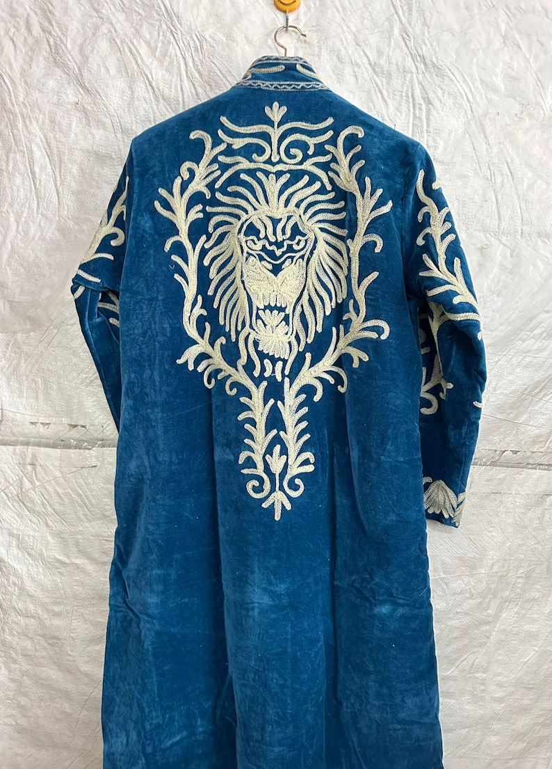 May include: A long-sleeved, deep blue velvet coat featuring intricate, cream-colored embroidery on the back. The design showcases a large, detailed lion head surrounded by ornate, swirling floral patterns that extend down the center of the garment.