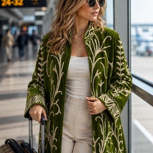May include: A long-sleeved olive green velvet coat featuring intricate cream-colored embroidery in a branching, organic pattern. The coat is worn over a white tank top and beige trousers, paired with round sunglasses and a gold pendant necklace.