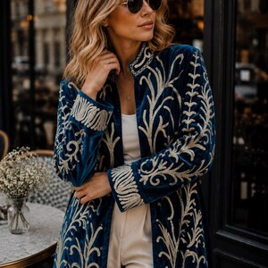 May include: A long-sleeved, deep blue velvet coat featuring intricate, light-colored embroidery in a swirling, organic pattern. The garment has a high collar, structured cuffs, and an open front design worn over a simple white top and light trousers.