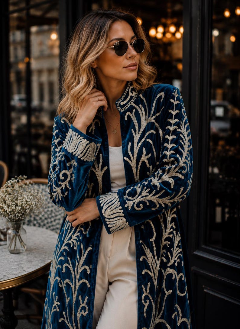 May include: A long-sleeved, deep blue velvet coat featuring intricate, light-colored embroidery in a swirling, organic pattern. The garment has a high collar, structured cuffs, and an open front design worn over a simple white top and light trousers.