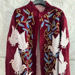 May include: A deep burgundy velvet coat featuring intricate embroidery of white doves and blue and gold leafy vines. The garment has a high collar, long sleeves, and an open front design, revealing a vertical red and white striped inner lining.