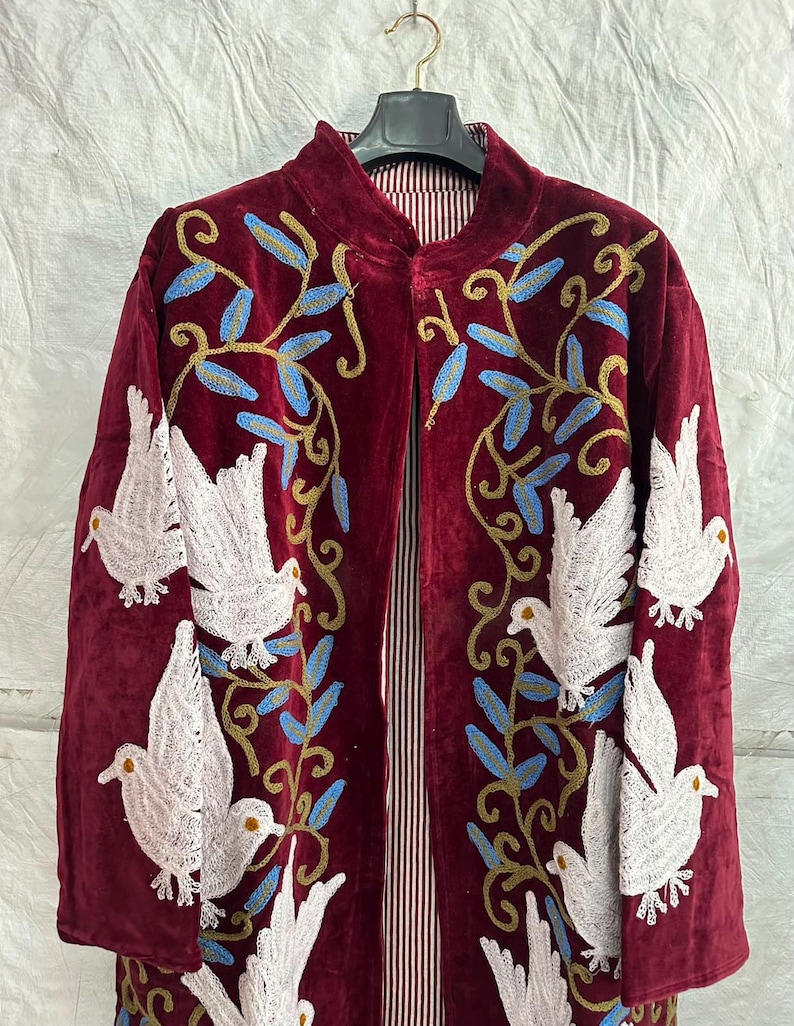 May include: A deep burgundy velvet coat featuring intricate embroidery of white doves and blue and gold leafy vines. The garment has a high collar, long sleeves, and an open front design, revealing a vertical red and white striped inner lining.