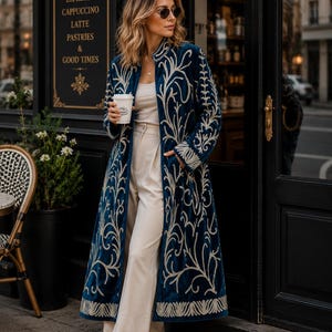 May include: A long, deep blue velvet coat featuring intricate cream-colored embroidery in a swirling, organic pattern along the front panels, cuffs, and collar, worn over a cream-colored top, cream trousers, and white sneakers.