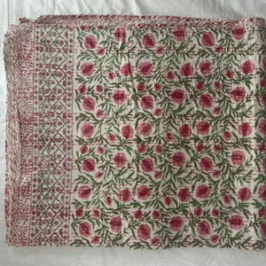 Könnte beinhalten: Ein rechteckiges Textil mit einem sich wiederholenden Muster aus rosa Blüten und grünen Blattreben auf cremefarbenem Hintergrund. Der Stoff enthält einen markanten vertikalen Rand mit einem geometrischen und floralen Design in Rosa- und Grüntönen.