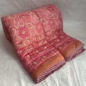 Puede incluir: Un edredón rosa y naranja doblado con un estampado floral. El edredón presenta un diseño repetitivo de grandes flores rosas sobre un fondo rosa más claro. El borde tiene rayas y detalles florales. El edredón está hecho de un material acolchado suave.