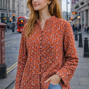 Op de afbeelding: Een shirt met lange mouwen en bloemenprint in oranje, rood en paars, met een opstaande kraag en knoopsluiting. Gedragen met een blauwe jeans. De achtergrond toont een stadsstraat met een rode bus en gebouwen.