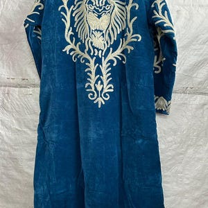 May include: A long-sleeved, floor-length blue velvet robe featuring intricate white embroidery. The back displays a large, detailed lion head design surrounded by ornate floral-like patterns, with additional decorative embroidery along the hem.