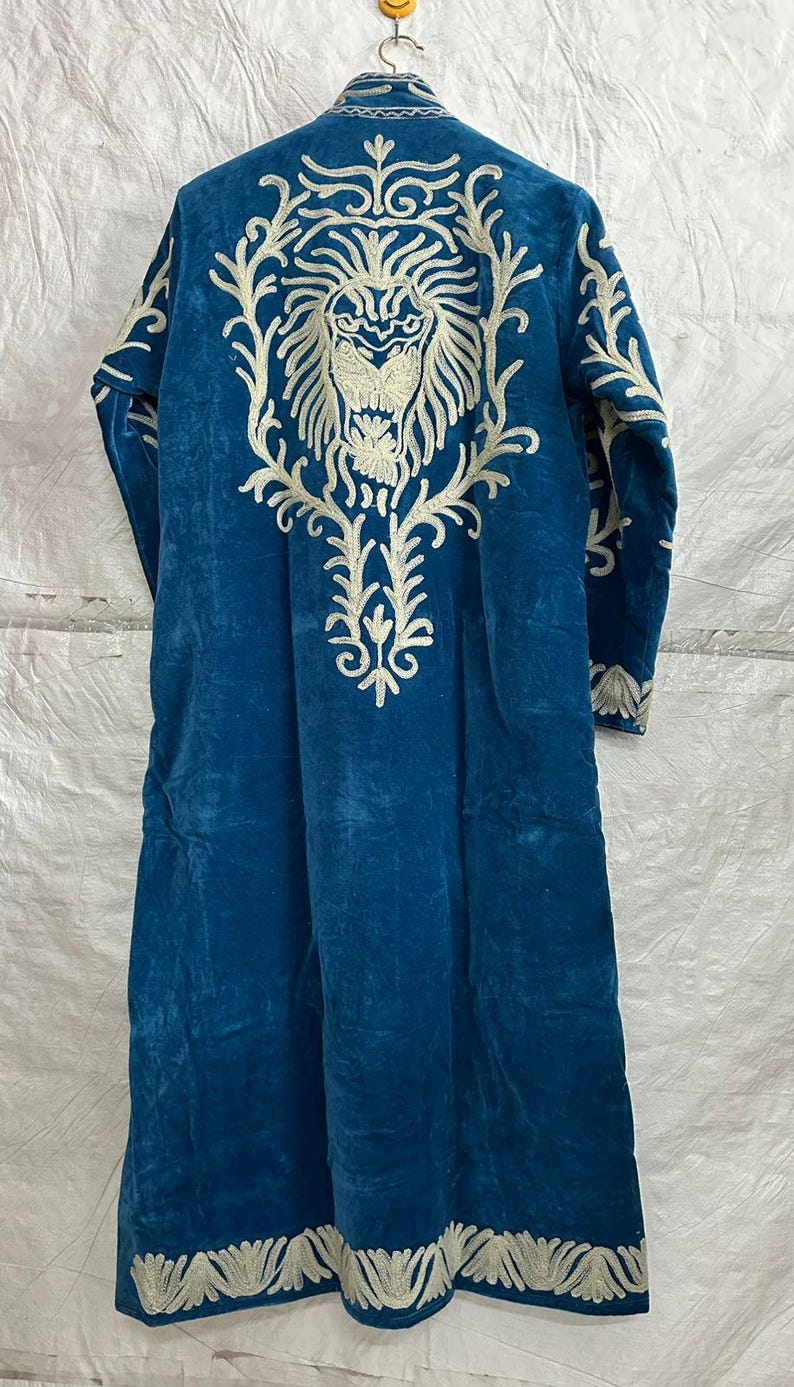 May include: A long-sleeved, floor-length blue velvet robe featuring intricate white embroidery. The back displays a large, detailed lion head design surrounded by ornate floral-like patterns, with additional decorative embroidery along the hem.