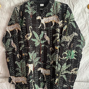 May include: A black long-sleeve quilted jacket featuring a repeating print of leopards, zebras, and tropical palm trees. The garment has a mandarin collar, a front button closure, and visible white running stitch details throughout the fabric.