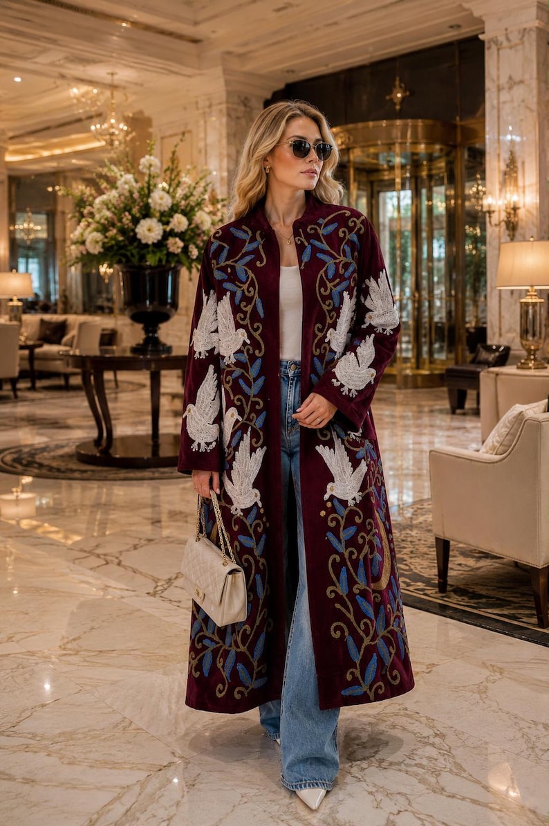 May include: A long, deep burgundy velvet coat featuring intricate white beaded bird embroidery and blue leaf patterns. The coat is worn over a white top and light blue denim jeans, paired with a cream-colored quilted handbag and white pointed-toe shoes.