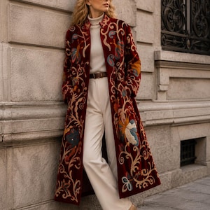 May include: A long, deep red coat featuring intricate, multi-colored floral and vine embroidery throughout. The coat is worn over a cream-colored turtleneck sweater, white trousers with a brown leather belt, and tan pointed-toe ankle boots.