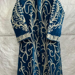 May include: A long-sleeved, knee-length coat in deep teal blue featuring intricate, light-colored embroidery in a branching, organic pattern that covers the front and sleeves. The coat has a high collar and a straight, open-front silhouette.