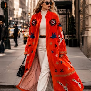May include: A long, vibrant orange coat featuring colorful embroidered motifs of suns, birds, and abstract shapes. The coat is worn over a white turtleneck sweater and white trousers, paired with a black handbag and black pointed-toe shoes.