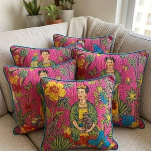 Op de afbeelding: Een collectie van vijf vierkante sierkussens met een felroze achtergrond en een herhalende print van een vrouw met donker haar, apen, papegaaien, vlinders en grote gele bloemen. Elk kussen is afgewerkt met een blauwgroene bies en een doorgestikte textuur.